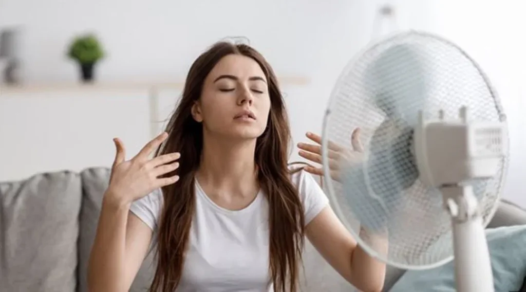 A woman cooling herself with a table fan while sitting on a couch.