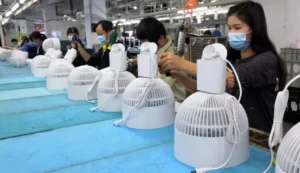 Workers on a factory assembly line wearing masks and assembling white electric fans. Multiple fan units are arranged on a blue workbench as employees use tools to complete the production process in a well-lit industrial setting.