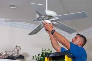 A repairman stands on a yellow stepladder to troubleshoot a white, multi-blade ceiling fan. His hands are raised and holding the central motor housing and light fixture, indicating he is performing an inspection or repair. This image represents a common home repair scenario for troubleshooting issues like a non-working fan, a wobble, or a broken light.