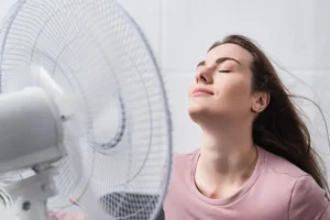 A young woman with long brown hair enjoys the cool breeze from a spinning electric fan, eyes closed and smiling with a relaxed expression, wearing a pink T-shirt in a bright indoor setting.