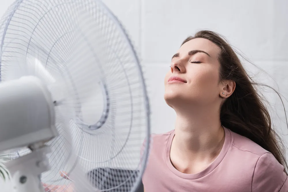 A young woman with long brown hair enjoys the cool breeze from a spinning electric fan, eyes closed and smiling with a relaxed expression, wearing a pink T-shirt in a bright indoor setting.