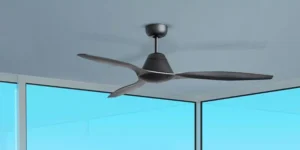 Modern black ceiling fan with three curved blades mounted on a sleek ceiling in a bright corner room with large glass windows and a clear blue sky outside.