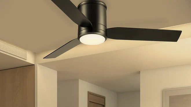 A low-angle shot of a low-profile (flush mount) ceiling fan with a sleek, matte black finish. The fan features three smooth blades and a central, integrated LED light which is turned on. It is installed on the ceiling of a modern, well-lit room, demonstrating a space-saving ventilation and lighting solution ideal for low ceilings.