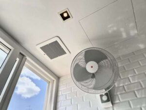 A modern bathroom ceiling featuring a square, built-in exhaust fan, a key component for a healthy bathroom. A separate wall-mounted fan is also visible, with a window showing a blue sky in the background.