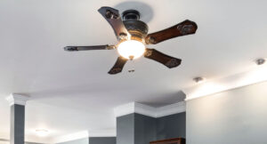 An upward-angle shot of a traditional, ornate ceiling fan with five dark wood blades and a bronze finish, installed on a flat white ceiling. The fan's central frosted glass light is turned on, and its pull chains are visible. The surrounding room has gray walls with white crown molding, illustrating a common home installation and raising the question of how high a ceiling fan should be for optimal airflow and safety.