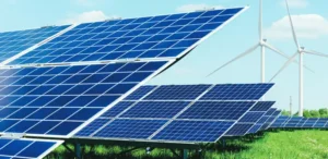 Rows of blue solar panels stretch across a vibrant green field under a clear blue sky, with two white wind turbines visible in the distance.