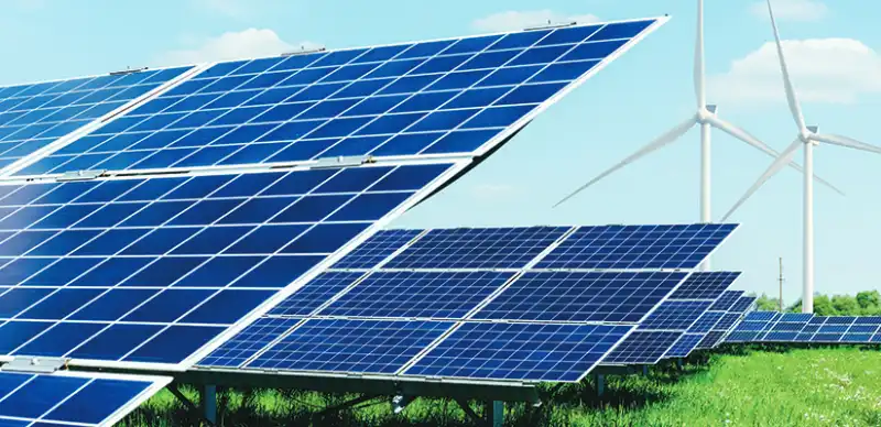 Rows of blue solar panels stretch across a vibrant green field under a clear blue sky, with two white wind turbines visible in the distance.
