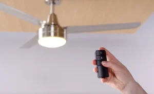 A close-up of a hand holding a black remote control and pressing a button to operate a modern ceiling fan. This image demonstrates how remote-controlled fans work, showing the fan with its light on and blades in motion against a wood-paneled ceiling.