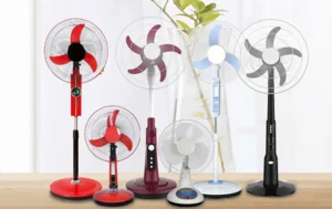 A lineup of various rechargeable fans in different sizes and colors, including red, black, blue, and white models, displayed on a wooden surface in front of a bright window with a potted plant in the background.
