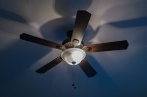 A five-blade wooden ceiling fan with an integrated light fixture casts distinct shadows on a blue ceiling, illustrating the comfort provided by ceiling fans and their evolution.