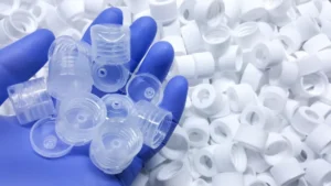 A hand wearing a blue glove holds a handful of clear ABS plastic disc top bottle caps. The background is filled with hundreds of similar opaque white ABS caps. The image illustrates the mass manufacturing process and quality control of injection-molded plastic components.