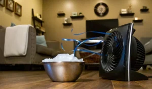 A small black fan blowing over a bowl of ice as a DIY air conditioner on a wooden living room floor.