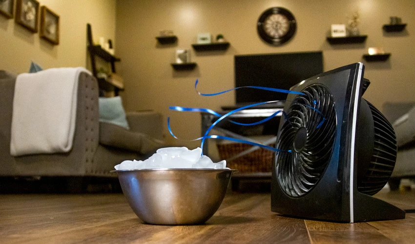 A small black fan blowing over a bowl of ice as a DIY air conditioner on a wooden living room floor.